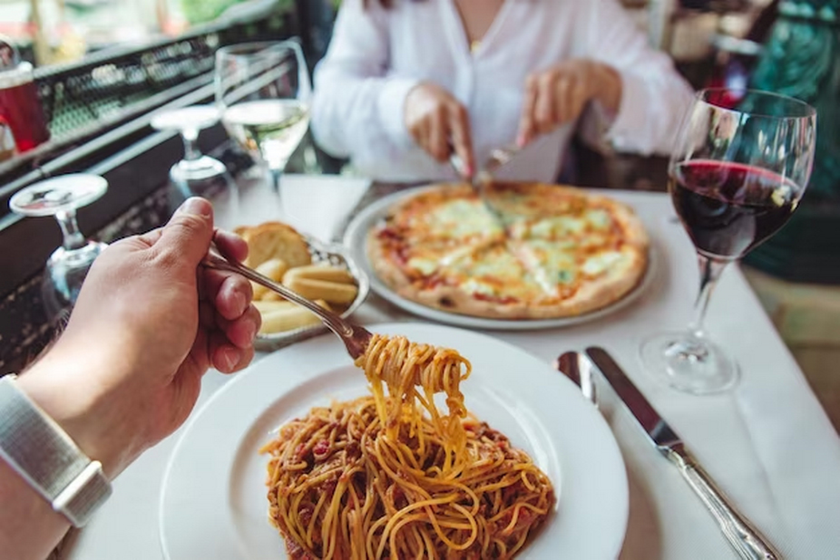 Person eating pasta with blurry woman slicing pizza in the background